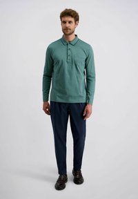 Mann trägt ein türkisfarbenes Langarm-Polohemd, marineblaue Hosen und dunkle braune Anzugschuhe und steht vor einem schlichten hellen Hintergrund.