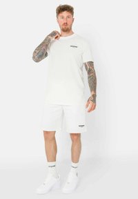 T-shirt bianco a maniche corte con testo nero, abbinato a pantaloni corti bianchi con un piccolo logo. Sneakers e calze bianche completano l'outfit.