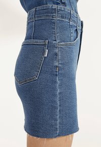 Nära bild av en person som bär en åtsittande blå denimkjol med en bakficka och en liten tygmärke, fransad fåll synlig.