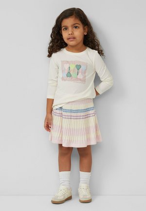 Jeune fille aux cheveux bouclés portant un haut à manches longues crème avec des motifs de cœurs et une jupe plissée à rayures pastel.