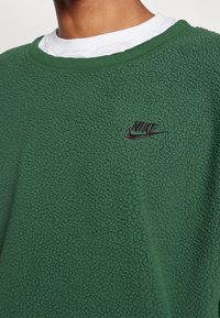 Zelená fleecová mikina s texturovaným povrchem, s kulatým výstřihem, bílou vnitřní vrstvou a černým vyšitým logem Nike na hrudi.