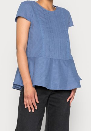 Blauwe blouse met korte mouwen, met getextureerde verticale lijnen en oogjesdetails, peplum zoom, gedragen met zwarte broek, getoond van nek tot heupen.