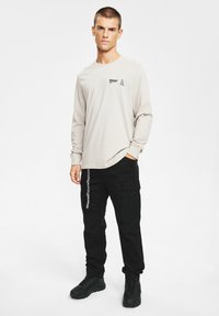 Beżowa koszula z długim rękawem z czarnym logo, zestawiona z czarnymi spodniami cargo z bocznymi kieszeniami i pionowym białym napisem. Czarne obuwie.