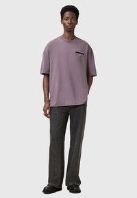 Camiseta oversized morada de manga corta, con un parche de logo negro en el pecho izquierdo. Combinada con unos vaqueros oscuros acampanados y zapatos negros.