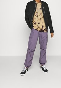 Chaqueta bomber negra, camiseta beige y marrón con efecto tie-dye, pantalones cargo morados y zapatillas negras y blancas con cierre de cordones.