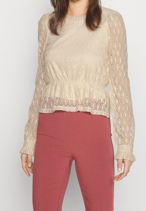 Blouse beige en dentelle à manches longues avec motifs circulaires et poignets à volants, associée à un pantalon taille haute rose poussiére.
