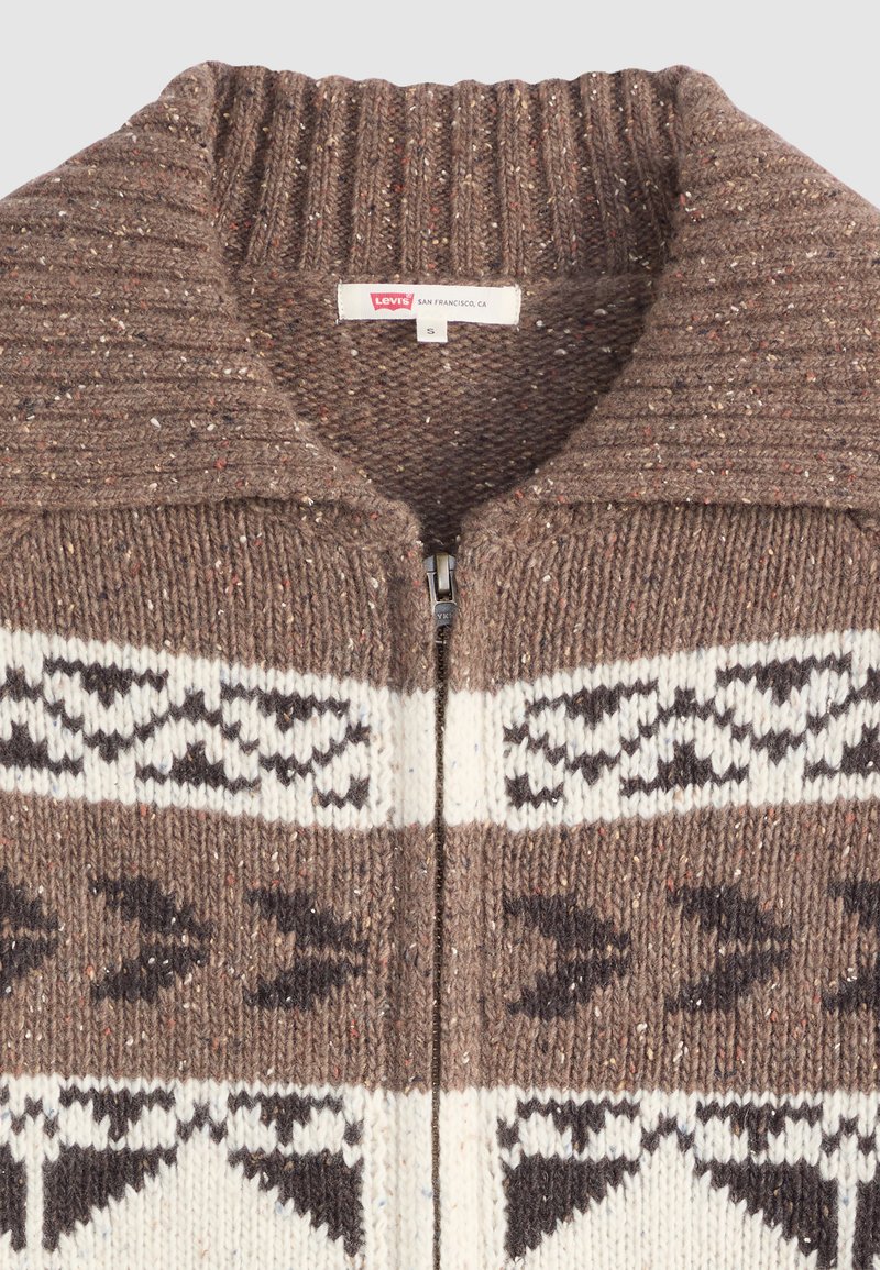 Brun strikket cardigan med høj ribkant, der har et geometrisk mønster i crème og mørkebrunt, lynlås-lukning og en spottet tekstur.