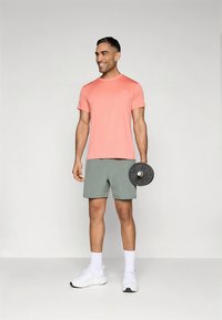 Ținută sport pentru bărbați: tricou roz cu mâneci scurte, pantaloni scurți verde deschis, ținând o placă de greutate neagră, cu adidași albi și șosete albe.