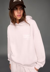 Svetlo ružová mikina s grafikou na prednej strane, mäkká látka, voľný strih a klokanie vrecko. Doplnok tmavohnedá šiltovka.