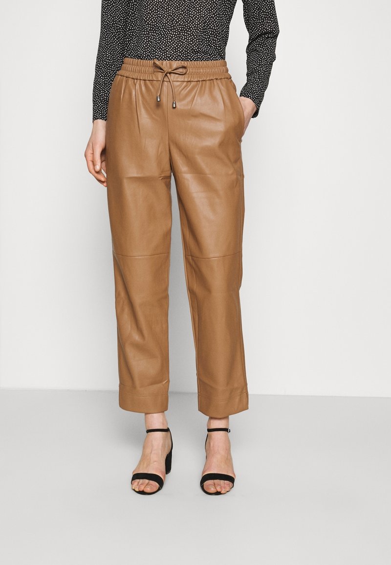 Femme portant un pantalon en cuir marron taille haute avec taille à cordon et des sandales noires à talons, debout devant un fond uni.