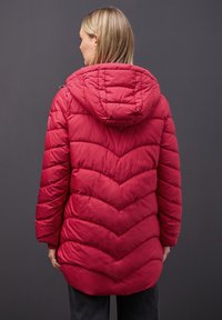 Femme aux cheveux blonds portant une veste d'hiver rouge matelassée avec capuche, vue de dos sur un fond sombre uni.