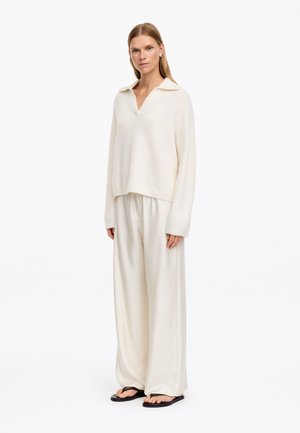 Femme aux cheveux blonds et raides portant un pull ample crème, un pantalon large crème et des tongs noires, debout devant un fond blanc.