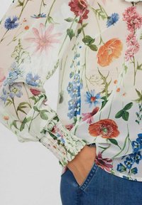 Genomskinlig blommig blus med en blandning av röda, blå och rosa blommor på en ljus bakgrund. Elastiska ärmslut och en avslappnad passform.