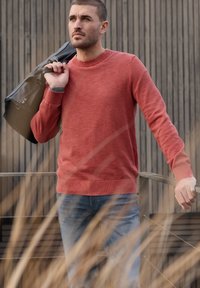 Homme en pull rouge et jean portant un sac noir sur l'épaule, debout en plein air devant un mur en bois avec de hautes herbes au premier plan.