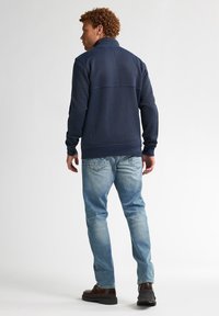 Sweatshirt din polar bleumarin cu guler înalt, țesătură texturată și manșete cu nervuri, purtat cu blugi de denim deschis și cizme maro, văzut din spate.