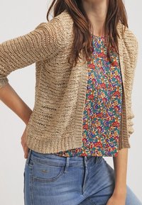 Lätt, beige stickad cardigan med öppen framsida, tillsammans med en livlig blommönstrad topp och blå denimjeans.