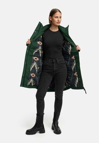 Groene buitenjas met een gestreepte zwarte voering. Model draagt een zwarte top en een donkere spijkerbroek, gecombineerd met zwarte enkellaarsjes.