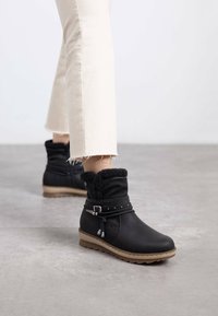 Botas negras hasta el tobillo con forro de piel sintética, una correa con acentos metálicos y una suela texturizada. Llevadas con jeans blancos cortos y deshilachados.