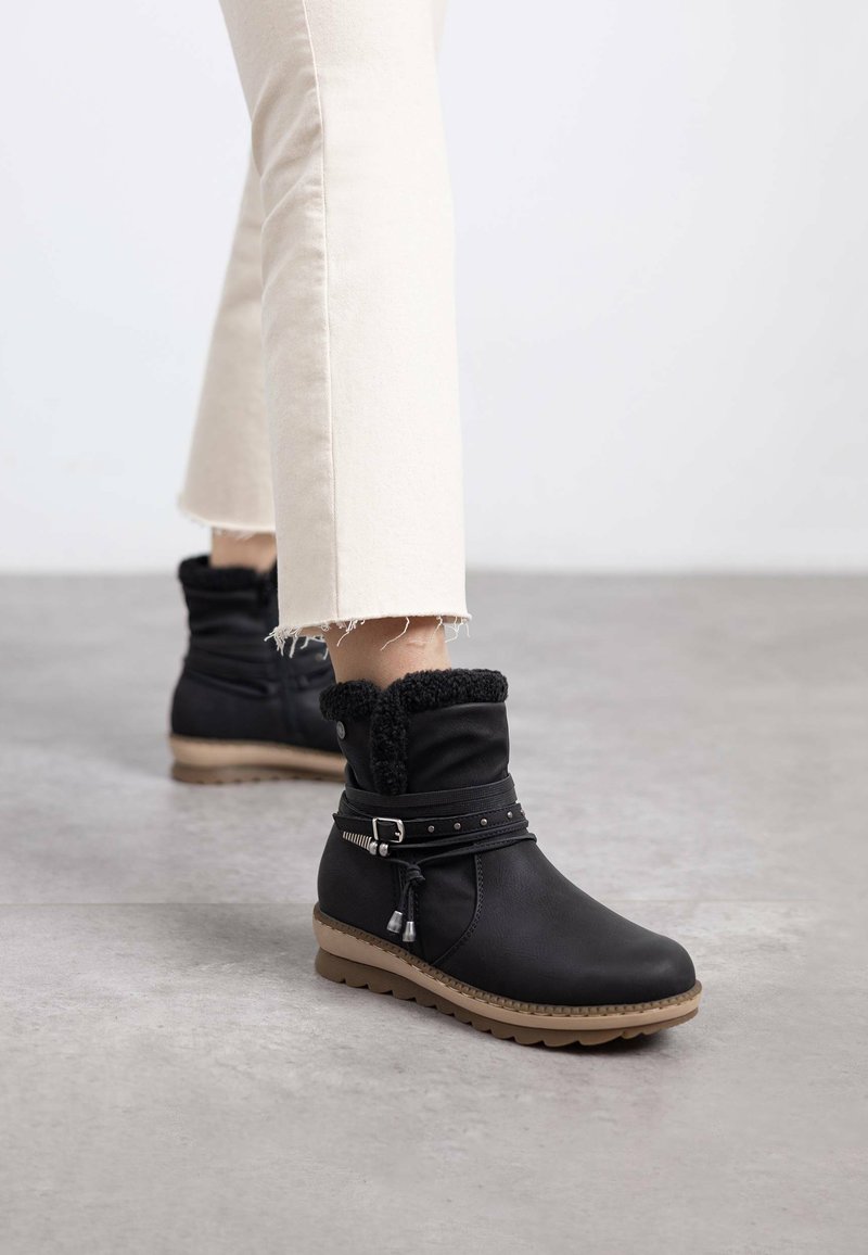 Botas negras hasta el tobillo con forro de piel sintética, una correa con acentos metálicos y una suela texturizada. Llevadas con jeans blancos cortos y deshilachados.