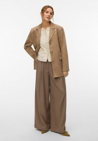 Femme portant un chemisier beige boutonné, un blazer ample couleur noisette, un pantalon large marron et des chaussures pointues couleur moutarde sur un fond uni.