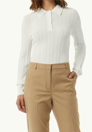 Polo blanc à manches longues côtelé avec boutons, associé à un pantalon beige taille haute et ajusté, une main dans la poche, fond neutre.