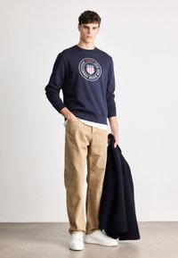 Granatowy sweter z okrągłym logo graficznym na piersi, w połączeniu z jasnobrązowymi spodniami i białymi trampkami, trzymający granatowy płaszcz.