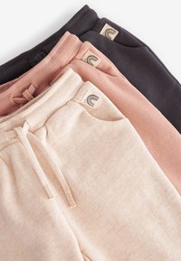 Trois paires de survêtements en beige, rose et noir. Chacune possède une taille côtelée, un cordon de serrage et une petite étiquette logo sur le côté.