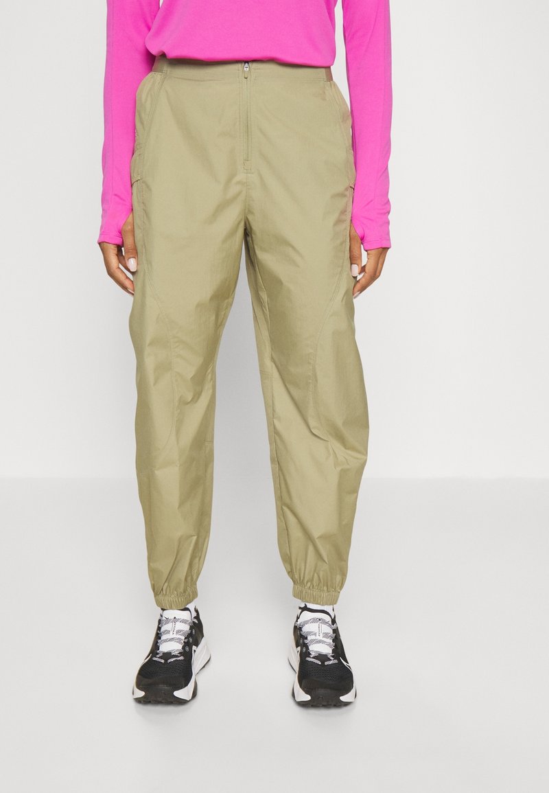 Personne portant un pantalon cargo beige avec une fermeture éclair à l'avant, des poignets élastiques, des baskets noires et blanches, et un haut à manches longues rose vif.