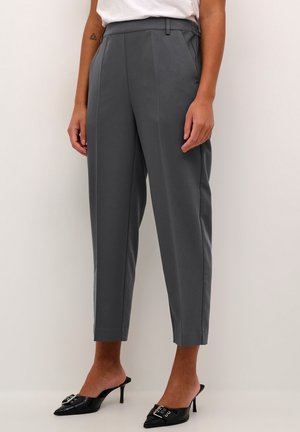 Persoon die cropped donkergrijze, op maat gemaakte broek met voorplooien draagt, witte top en zwarte pumps met spitse neus en hakken met gespsluiting.