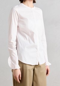 Femme portant un chemisier blanc à manches longues avec des poignets volants et un pantalon beige, debout devant un fond gris clair uni.