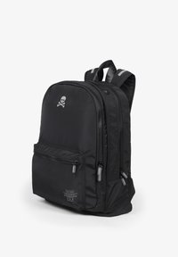 Mochila negra de nailon con múltiples compartimentos con cremallera, destacando un logotipo de calavera en la parte frontal y correas de hombro a rayas. Textura suave.