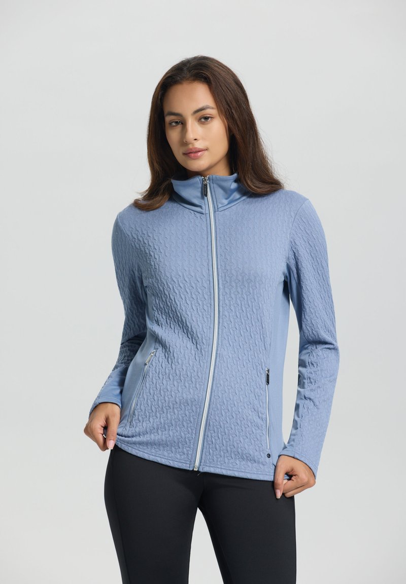 Lyseblå lynlåsjakke med et struktureret mønster, høj krave og sidelommer, sammen med sorte leggings.