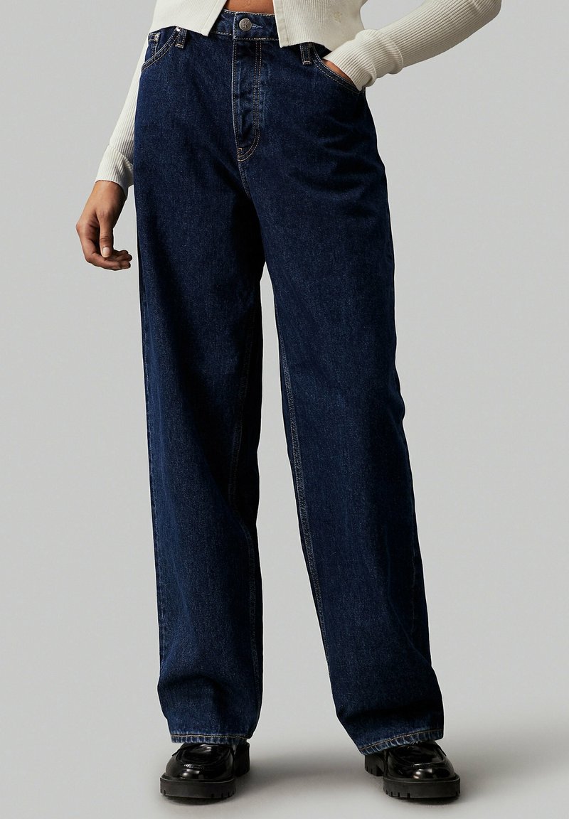 Donkerblauwe denimbroek met een wijde pasvorm, hoge taille, standaardzakken en contrasterende stiksels. Bevat riemlussen en een ontspannen snit.