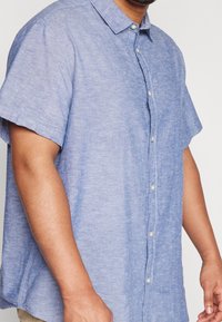 Světle modrá košile na krátký rukáv s límečkem, vyrobená z texturované látky. Na přední léze jsou viditelné knoflíky.