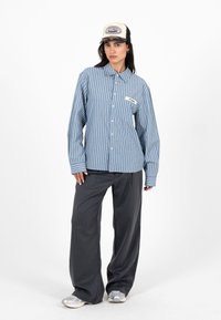 Femme portant une chemise à rayures bleues boutonnée, un pantalon ample gris foncé, des baskets et une casquette de camionneur blanche et noire, debout devant un fond blanc.