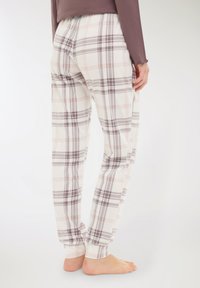 Persona a piedi nudi con pantaloni della tuta a quadri bianchi e beige e una maglietta a maniche lunghe color mauve, in piedi contro uno sfondo chiaro e uniforme.