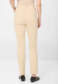 Pantalons skinny beiges avec poches arrière, fabriqués en tissu extensible. Portés avec des ballerines noires. Présentent une taille ajustée et une conception longueur cheville.