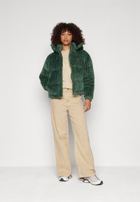Groene corduroy pufferjack met ribbeltextuur, ritsluiting en hoge kraag. Beiggekleurde T-shirt en beige wijde broek. Witte sneakers met accenten.