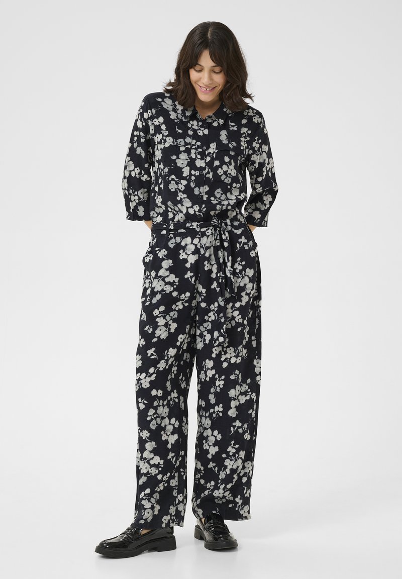 Mono floral en azul marino con estampados blancos, con un cuello con solapa, mangas de tres cuartos, cinturilla con lazo y pantalones de pierna ancha.