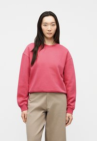 Femme aux longs cheveux noirs portant un sweat-shirt rose à col rond et un pantalon beige, debout devant un fond gris clair uni.
