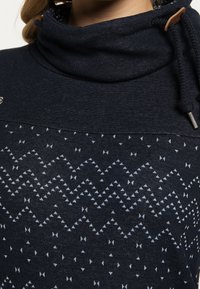 Tummansininen collegepaita korkealla kauluksella, jossa on kevyesti sinisistä geometrisista muodoista koostuva tekstuuripinta. Nyöri puuhengellä.