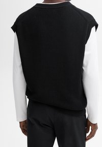 Schwarzer, gestrickter ärmelloser Pullover mit Rippstruktur und Rundhalsausschnitt. Wird über einem weißen Langarmshirt getragen, das über herunterhängende Schulternähte verfügt.