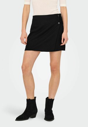 Falda envolvente negra de longitud media por encima del muslo, hecha de una tela suave, que presenta un lazo en el lado y dos botones. Combinada con botas de tobillo negras con textura.