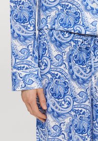 Kék és fehér paisley mintázatú szövet, bonyolult virágmintákkal. Hosszú ujjak gombos mandzsettával, sima textúrával.