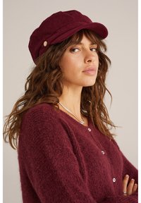 Burgundy kordflöjt keps med en strukturerad skärm och dekorativa sidoknappar, buren med en matchande texturerad cardigan med vita knappar.
