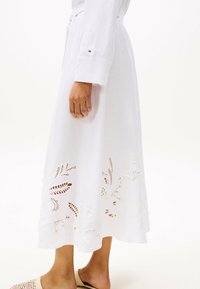 Vestido blanco de manga larga, con un intrincado diseño floral recortado cerca del dobladillo. Tela ligera y detalle de cintura anudada.