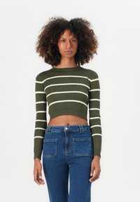 Olivengrønn kort genser med hvite horisontale striper, ribbestrikket tekstur og lange ermer, kombinert med høyhalsede blå jeans.