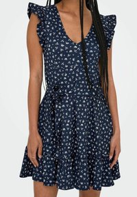 Marineblaues Kleid mit weißem Blumenmuster, Rüschensäumen, V-Ausschnitt und verstellbarem Taillengürtel. Stufenrock aus strukturiertem Material.