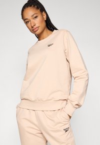 Hanorac bej deschis din material moale, cu decolteu rotund, manșete drepte și un mic logo negru Reebok pe piept.