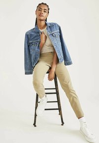 Giacca di jeans, t-shirt beige e chinos color cammello indossati con sneakers bianche. Modello seduto su uno sgabello di legno, sfondo neutro.